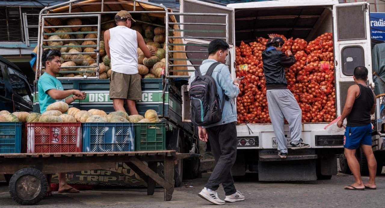 Men unloading vegetables from trucks at a bustling outdoor market.